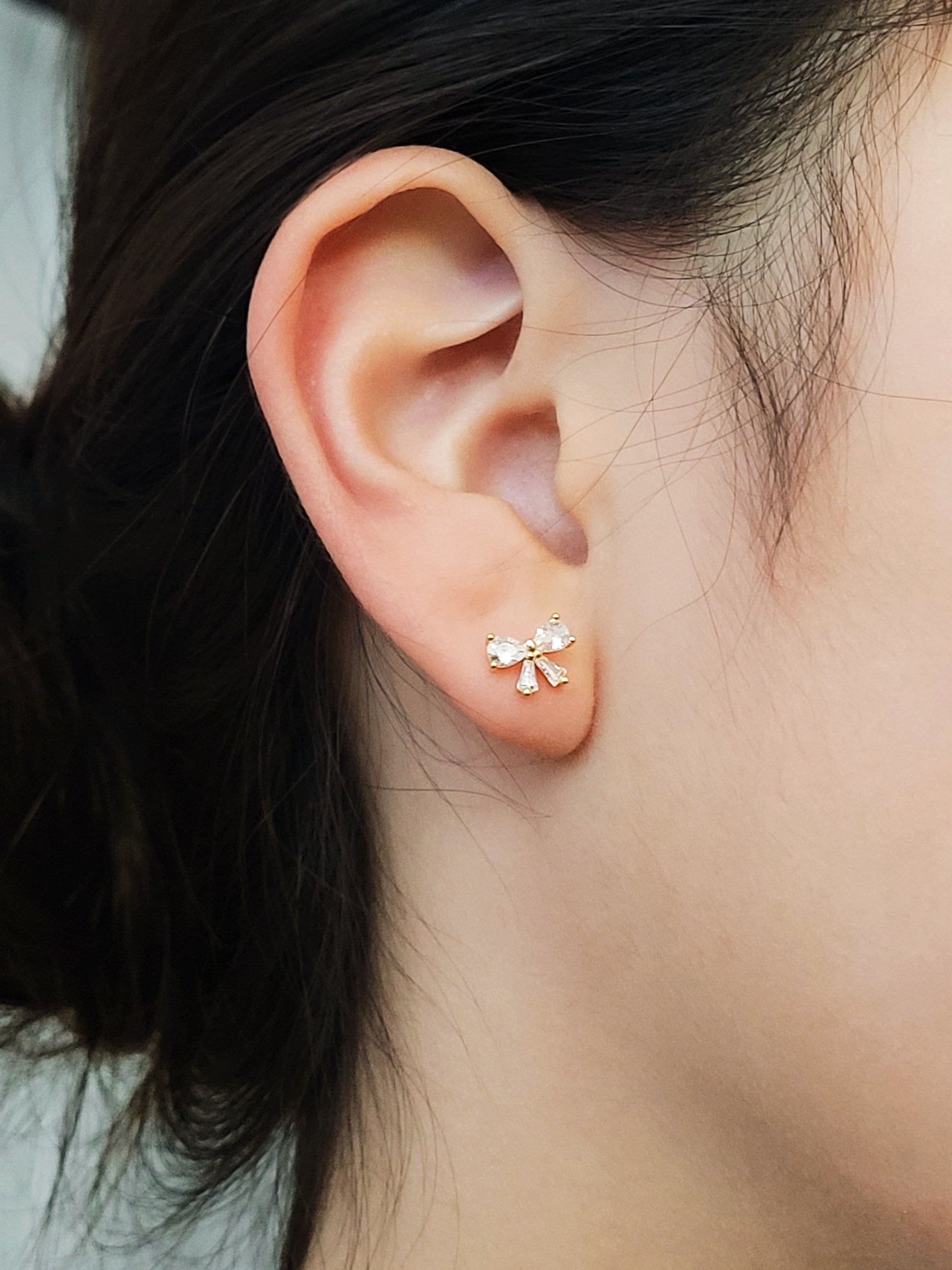 Close up of a woman's ear wearing dainty bow stud earrings with sparkling cubic zirconia and gold plating.