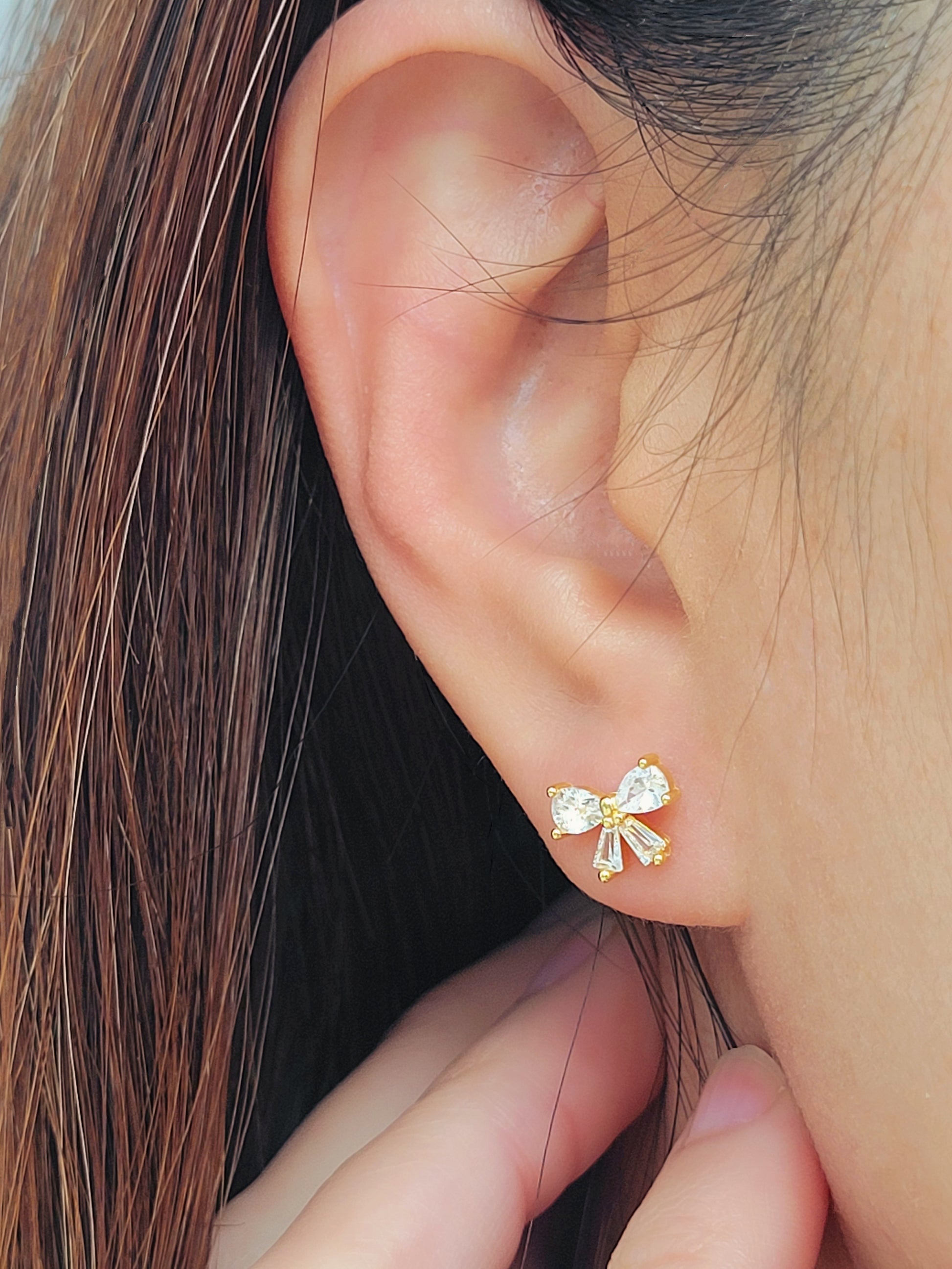 Close up of a woman's ear wearing dainty bow stud earrings with sparkling cubic zirconia and gold plating.
