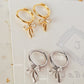 2 Pairs of Gold-plated and sterling silver ribbon bow dangly huggie earrings displayed on a neutral stone background.