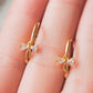 A pair of dainty gold plated ribbon bow dangly huggie earrings displayed on a woman's hand.