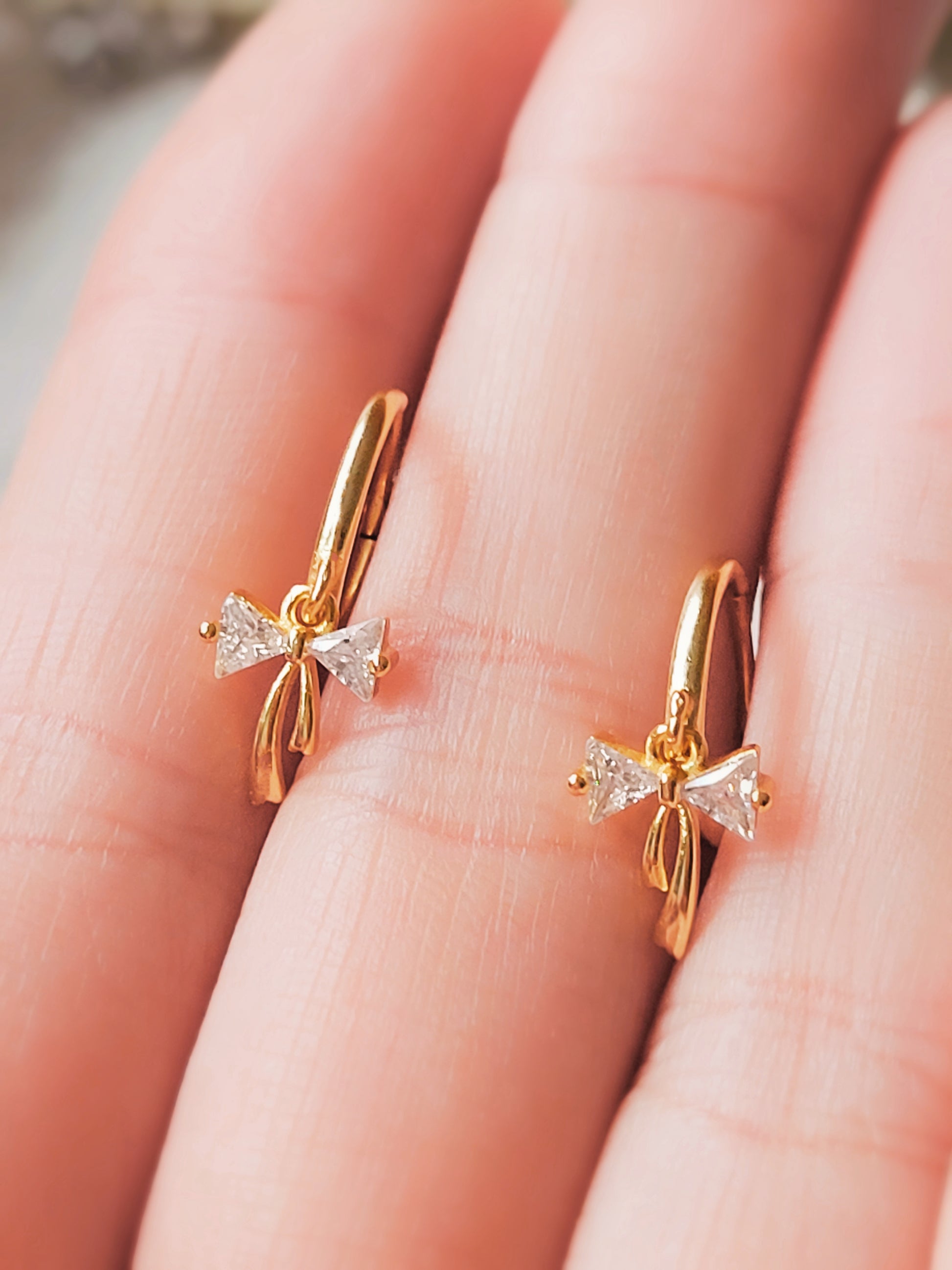 A pair of dainty gold plated ribbon bow dangly huggie earrings displayed on a woman's hand.