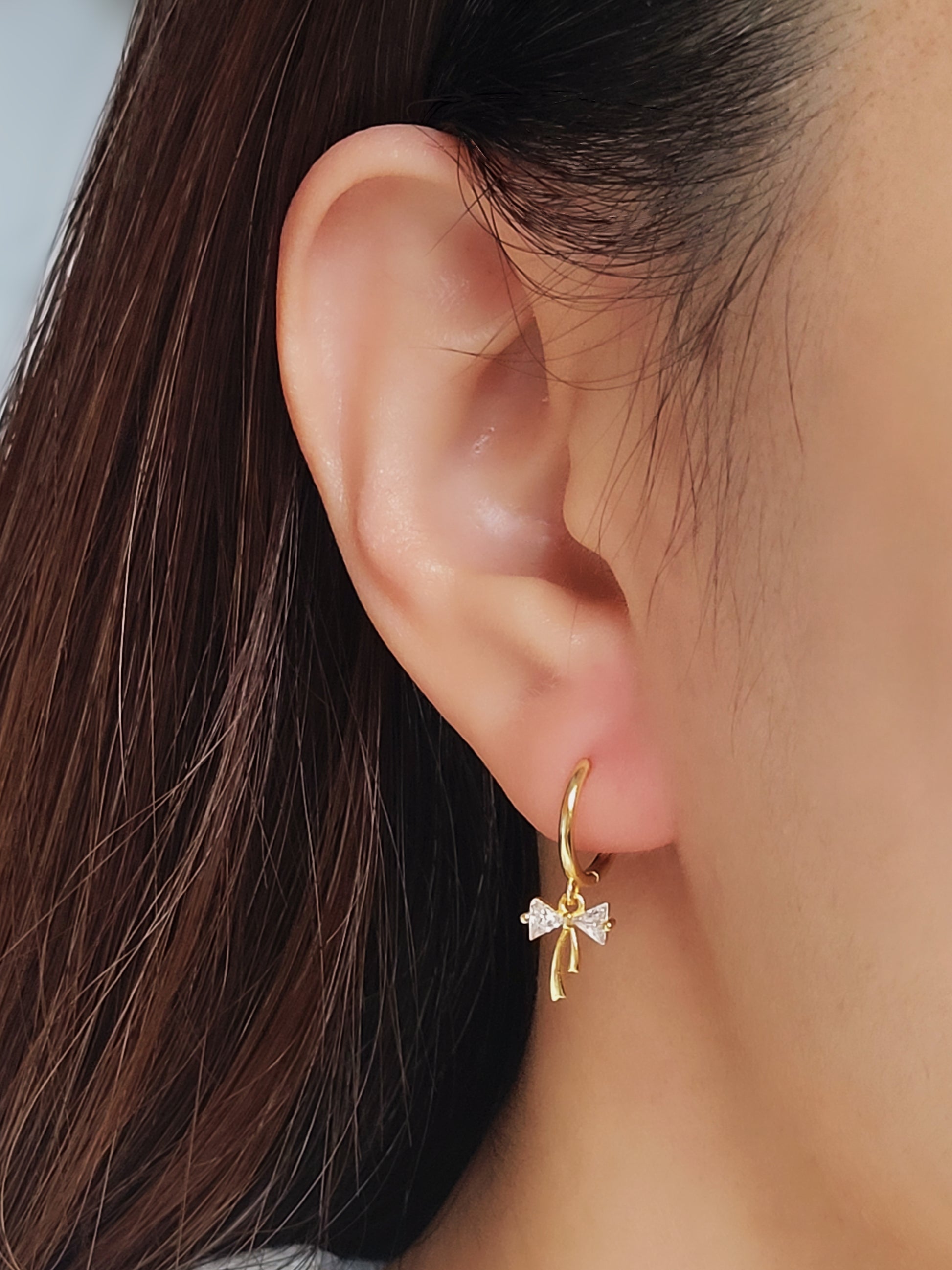 Close-up view of a woman's ear wearing gold ribbon bow earring with a dangling design, perfect for gifting to girlfriends, daughters, or loved ones.