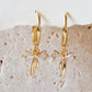 A pair of dainty gold plated ribbon bow dangly huggie earrings displayed on a neutral stone background.