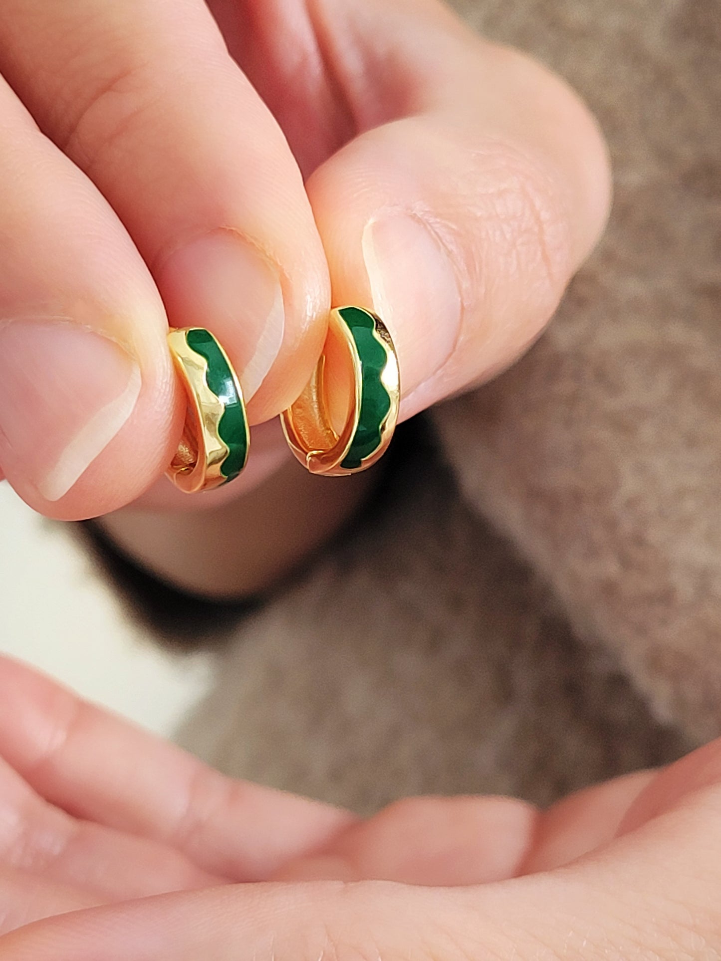 Gold and green enamel earrings held between fingers against a neutral background