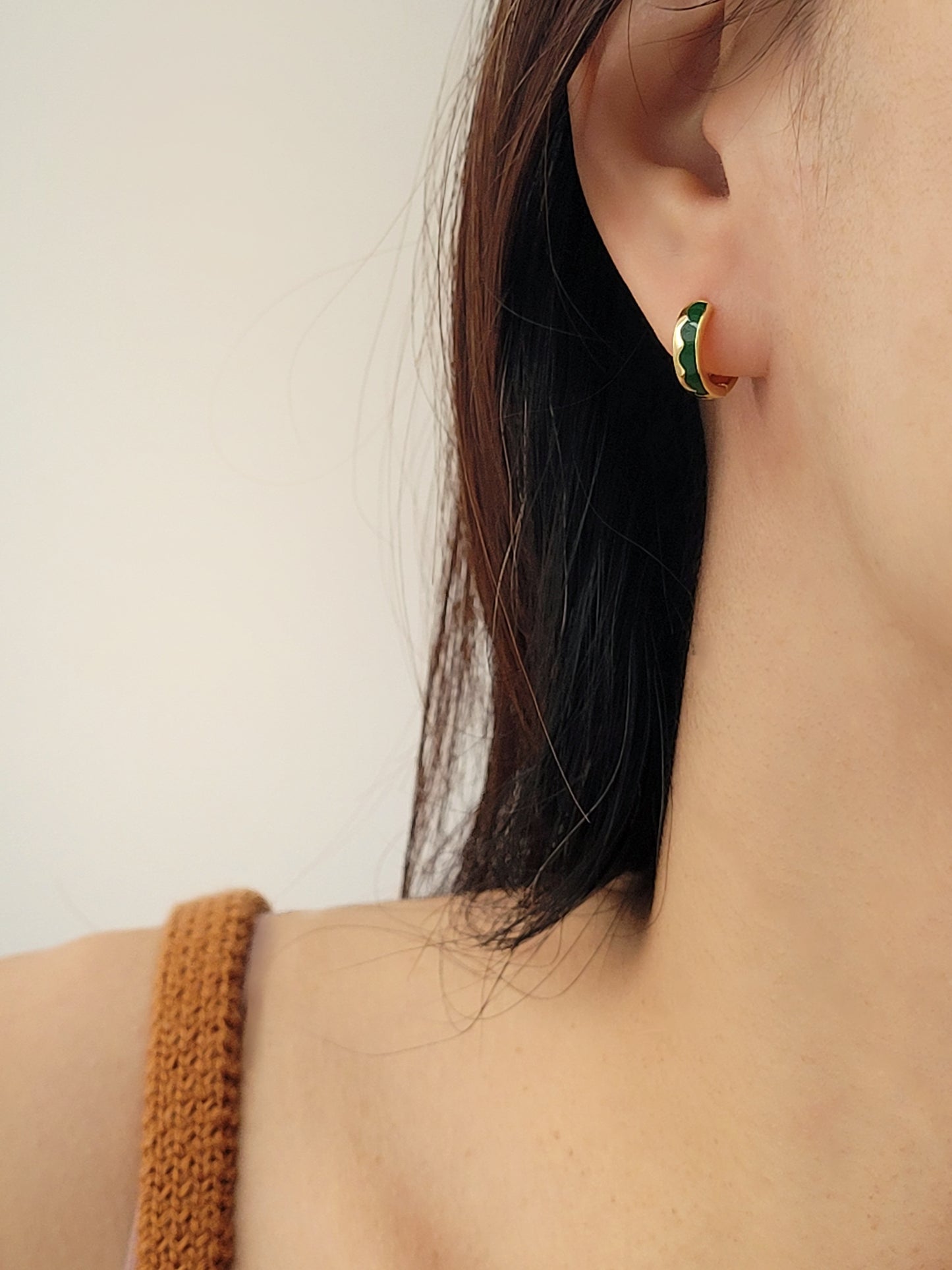 Close-up of an ear wearing a gold huggie hoop earring with green enamel on a neutral background