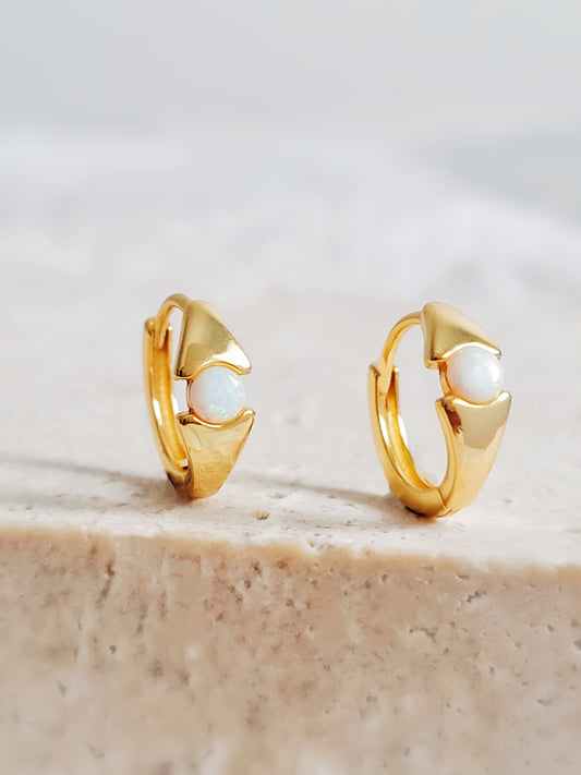 A pair of gold huggie hoop earrings with opal stones in evil eye design, displayed on a beige surface.