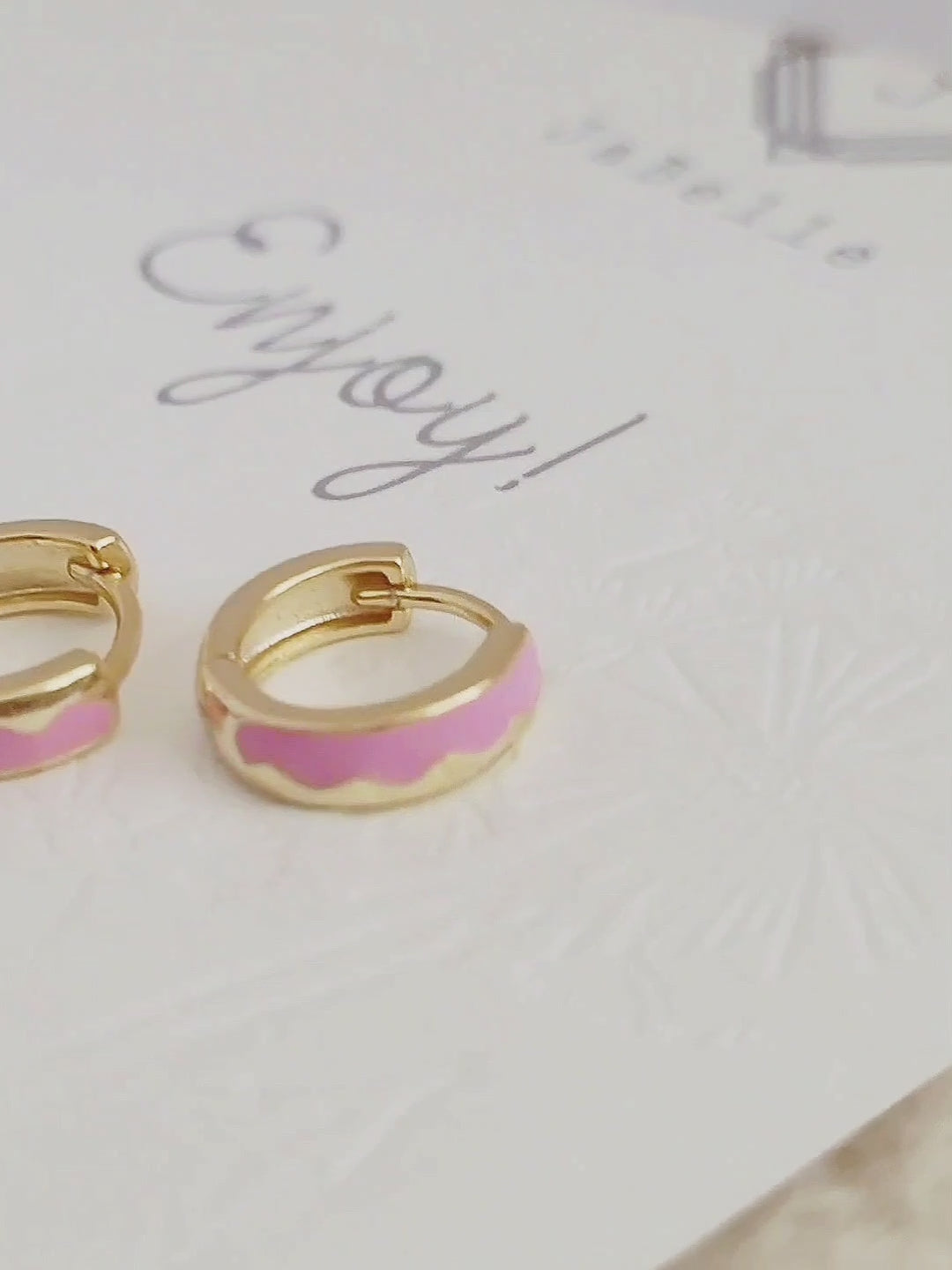 Details of a pair of chic pink enamel huggie hoops with a unique wavy pattern in 18K gold plating, perfect for adding a pop of color.