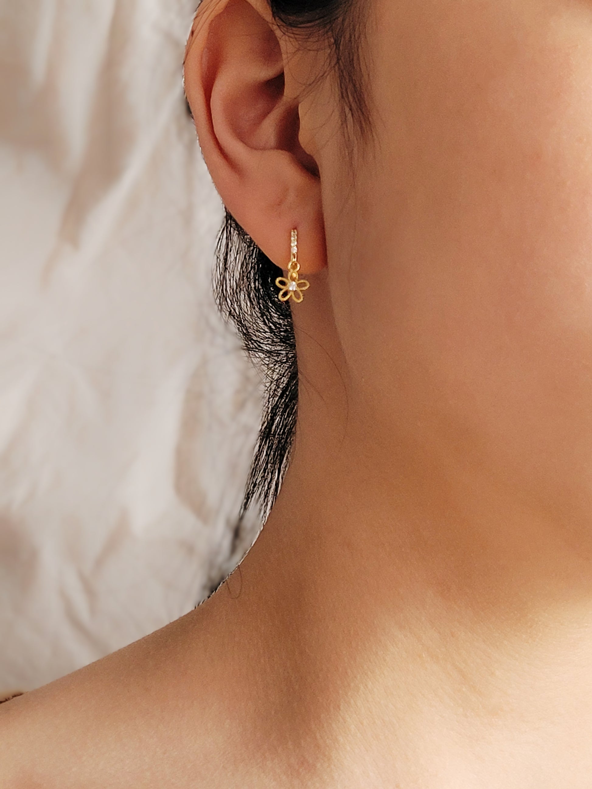 A close-up of a gold huggie earring with a delicate dangling flower charm, crafted in gold plated sterling silver. The intricate flower design adds a touch of femininity, making them ideal as wedding jewelry, a bridesmaid gift, or a thoughtful present for her.