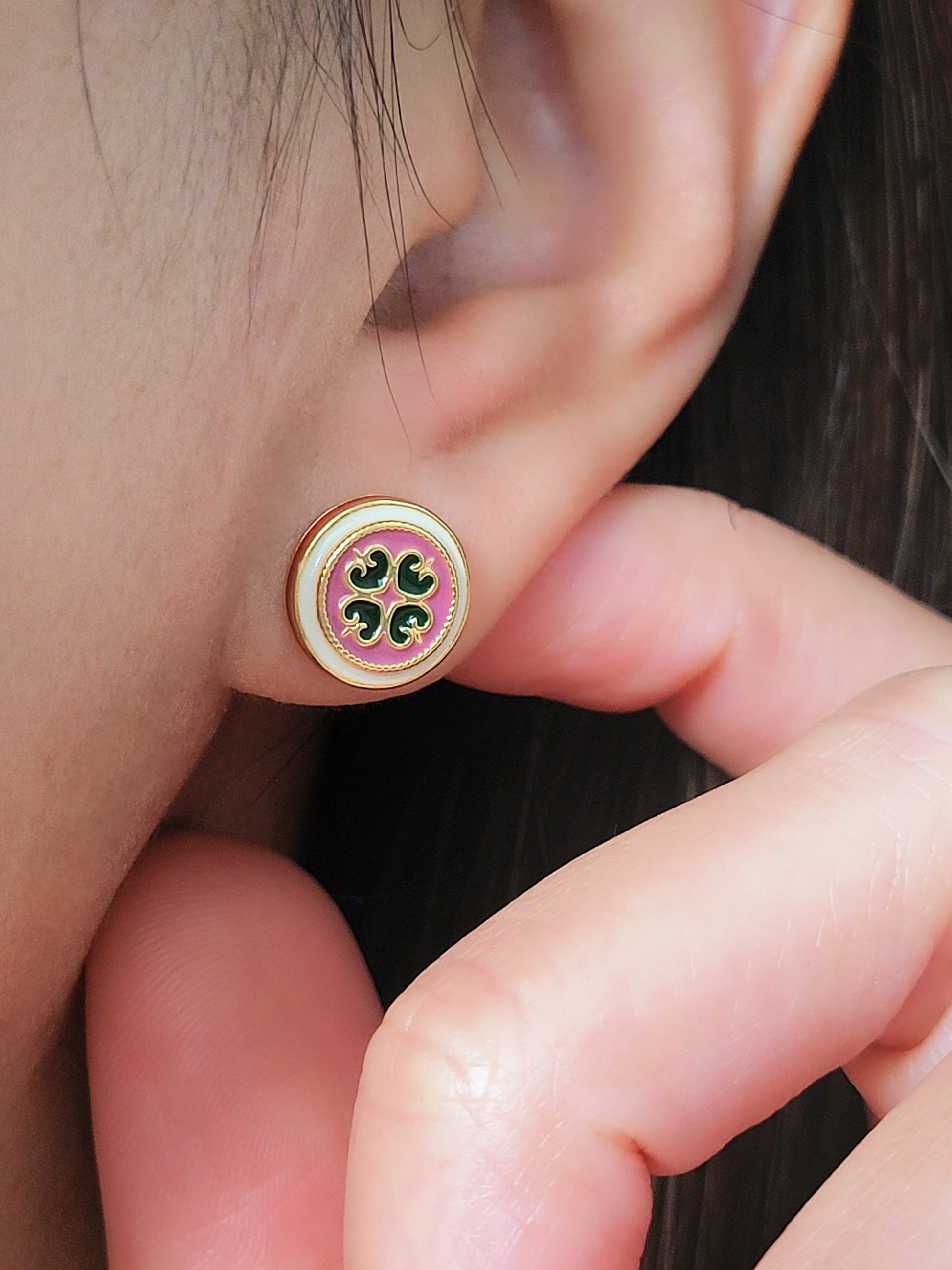 Close-up of a woman's ear wearing a pair of Retro Gothic tile pattern enamel stud earrings in pink, dark green, and gold. The intricate design features a vintage-style pattern with a bold color combination, creating a striking and elegant look.